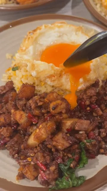 A spoon breaking into a crispy fried egg, the orange yolk running over minced-pork krapow and fried rice at King of KraPOW
