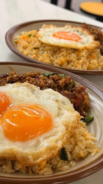 Two plates of King of KraPOW krapow over fried rice on a food court table, one with a double yolk